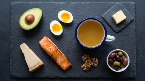 A mug of bone broth surrounded by keto foods including avocado, eggs, salmon, and cheese